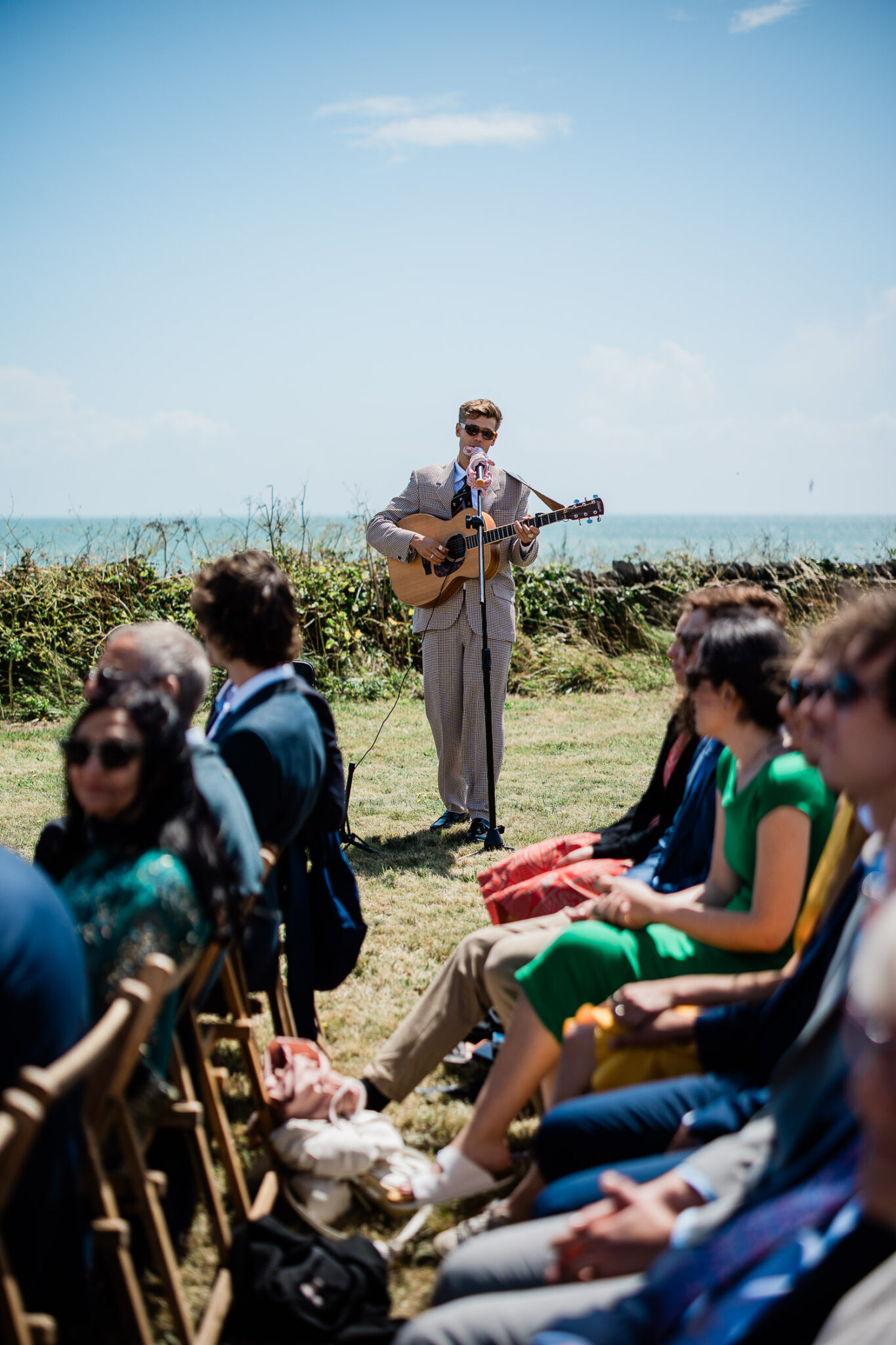 A guitar player sings acoustic covers whilst guests are arriving to outside wedding ceremony in Devon.