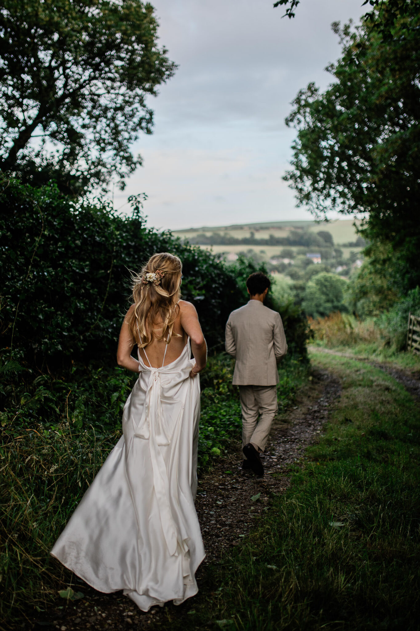 A bride and groom run down an English country lane, during a couples portrait session at a barn wedding venue in Devon, UK.