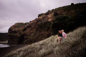 A couple share an intimate moment on a grassy hill at a picturesque black sand beach near Auckland