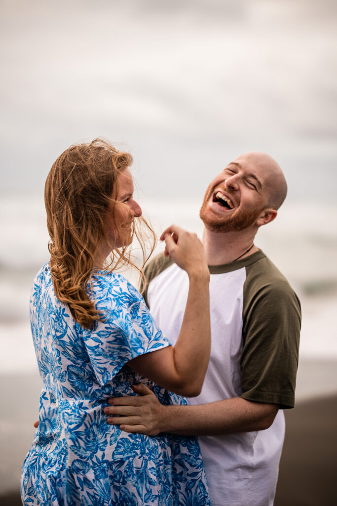 A young couple laugh together, during a documentary photography photoshoot at a beach in New Zealand