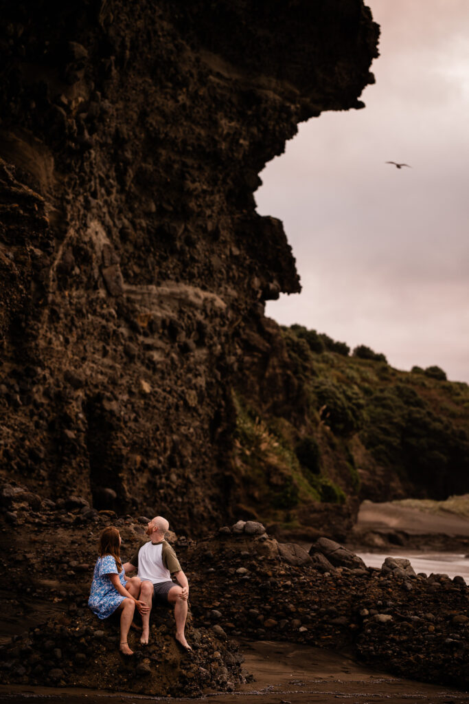 A couple sit under an impressive rock formation during a beach pre-wedding shoot in NZ
