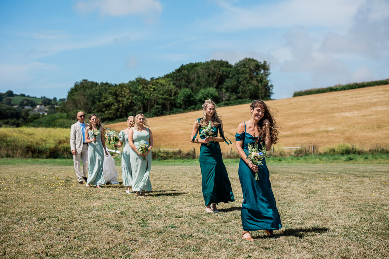 A bridal party walk through a field on their way to a bohemian ceremony at The Grain Store in Devon, UK.