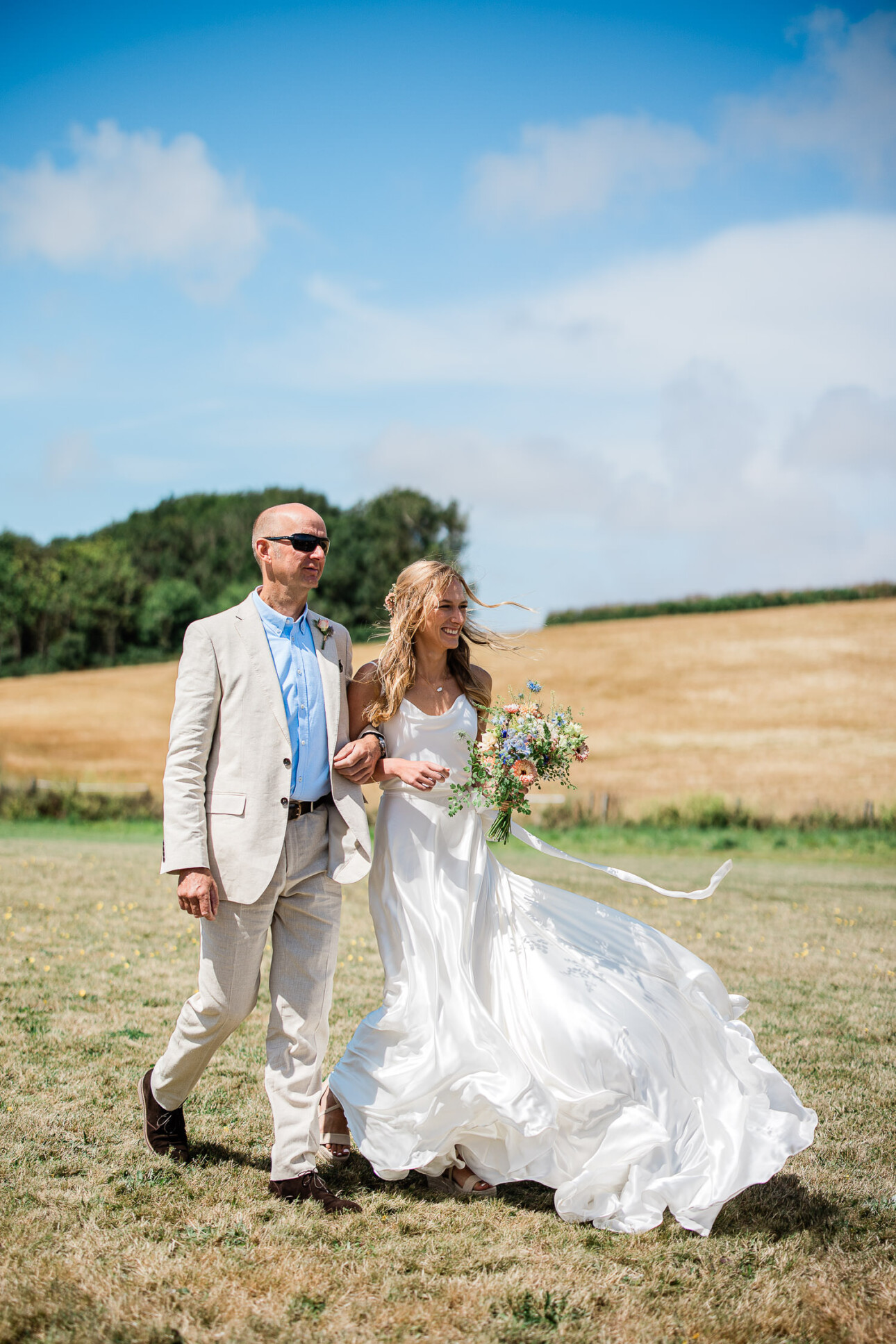 A bride and the father of the bride walk down the aisle of an open air ceremony in the South West.