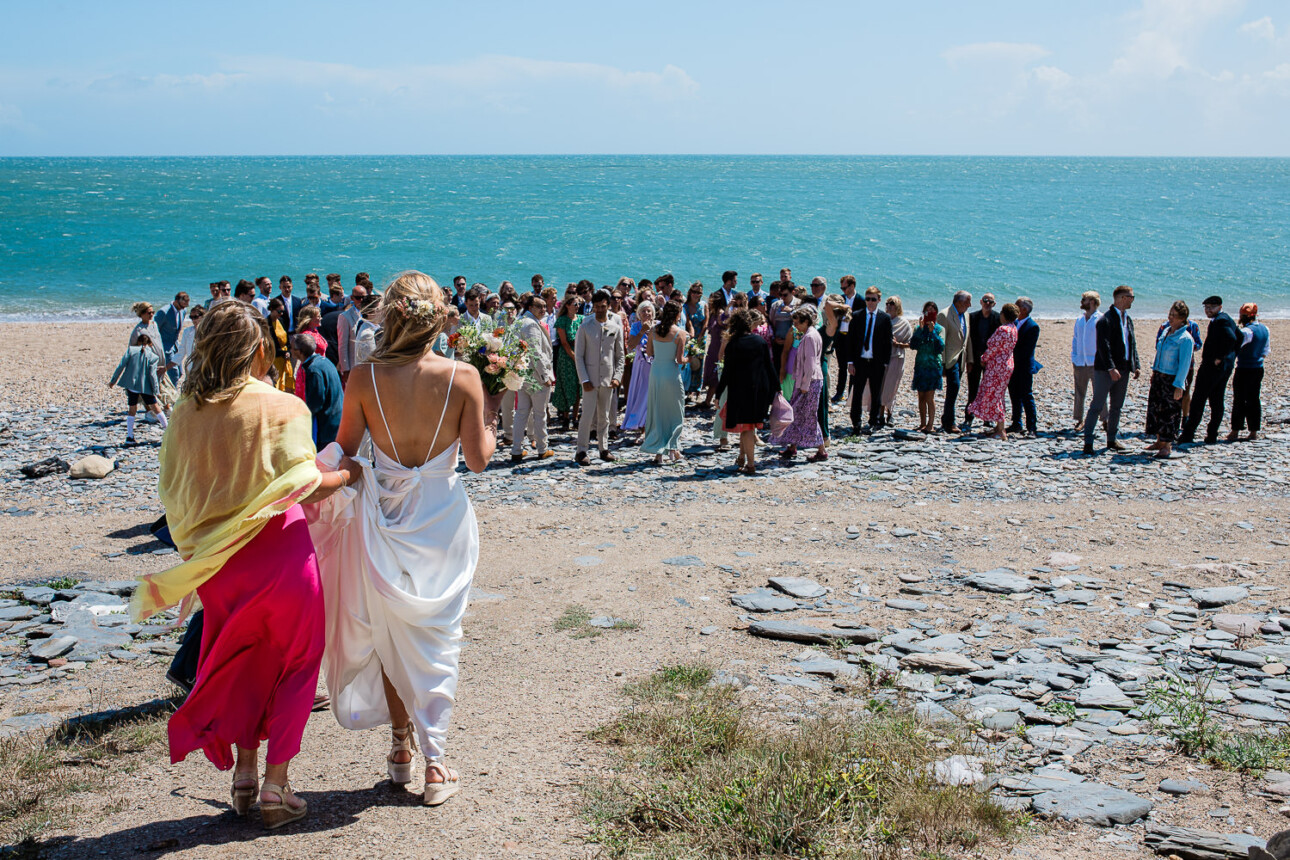 A bride and her friend walk down to join wedding guests on the beach in Devon, for a formal group photo of all the wedding guests.