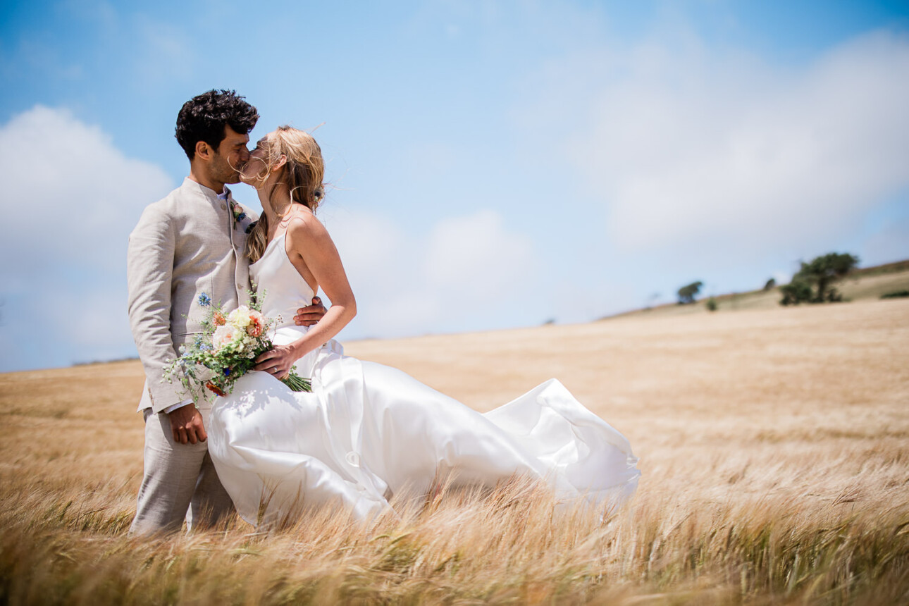 A wedding couple share a kiss in a barley field, during a quiet moment away from their outdoor bohemian inspired wedding in the UK.