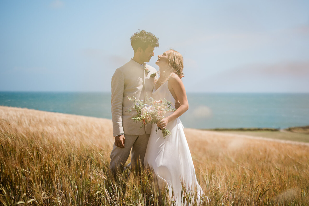 A couple smile at each other, during a couples portrait session in a field at The Grain Store wedding venue.