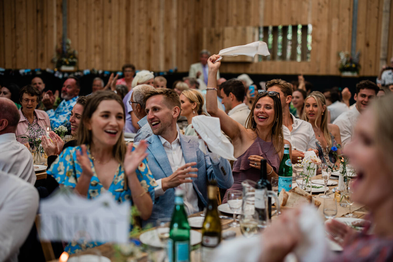Wedding guests wave napkins as the bride and groom enter their boho themed wedding reception in a barn wedding venue.