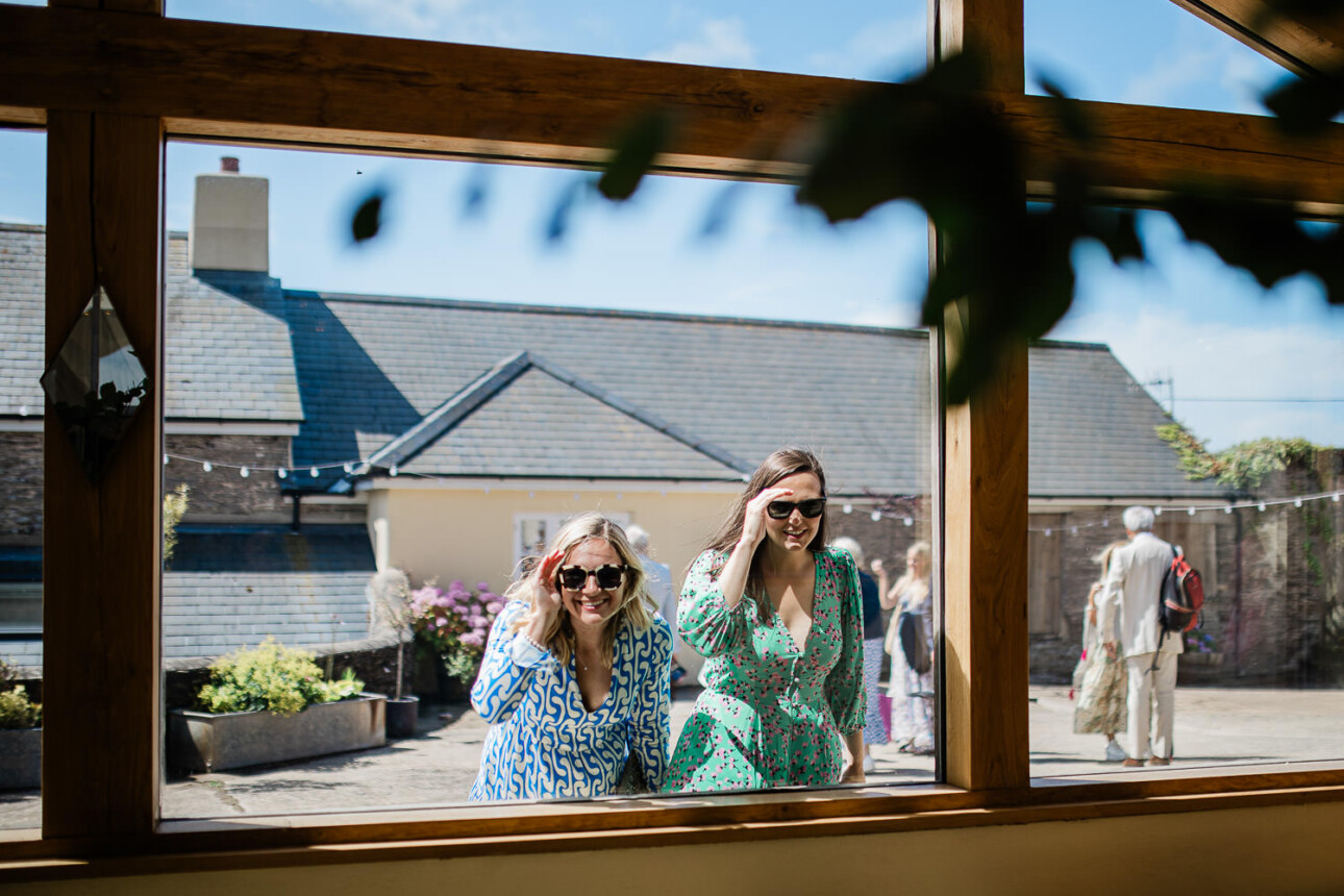 A documentary photography moment of two women looking through the window, into the wedding reception barn.