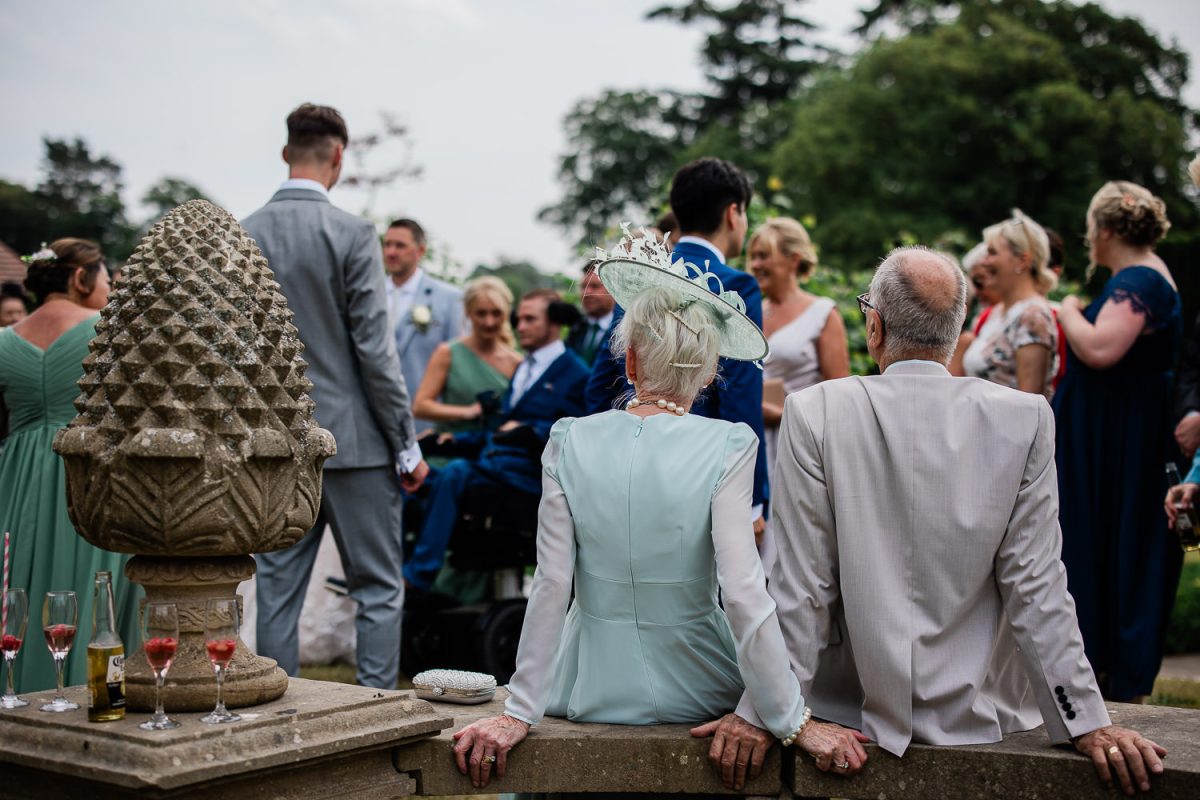 A candid capture of an elderly couple sit back and watch the wedding guests preparing for formal photos.