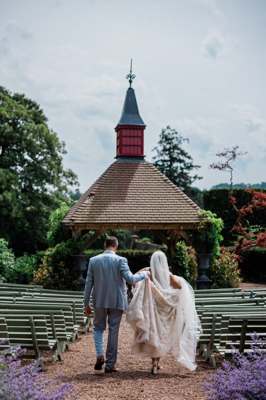 A groom helps the bride to hold her dress, as they walk down to a rotunda in English gardens.