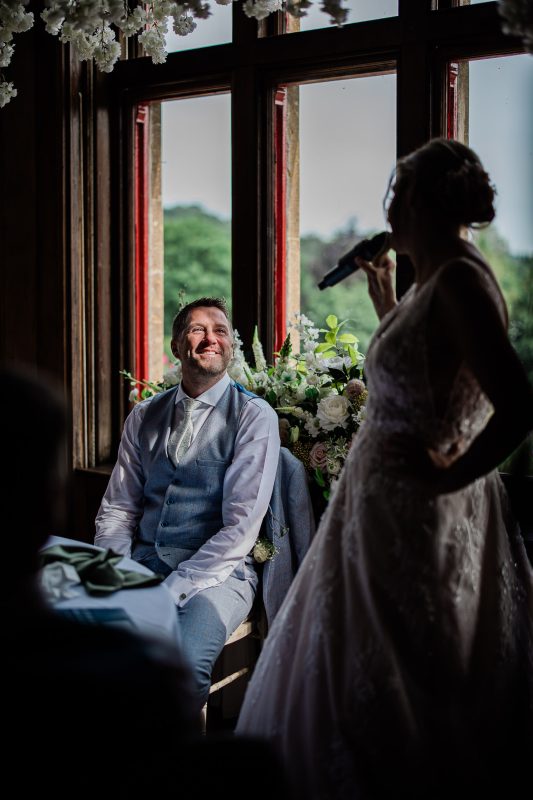 A groom smiles up at a bride, during her speech.