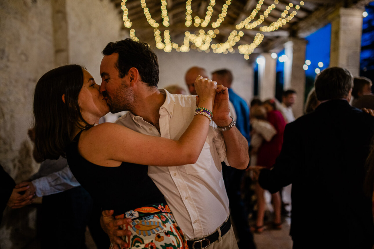 A couple lean in for a spontaneous kiss, during a wedding party.