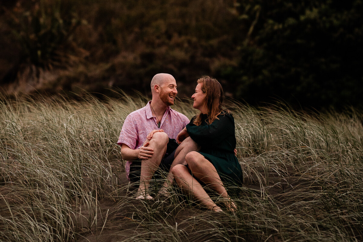 A newly engaged couple laugh together during a pre-wedding shoot at Piha by a documentary wedding photographer