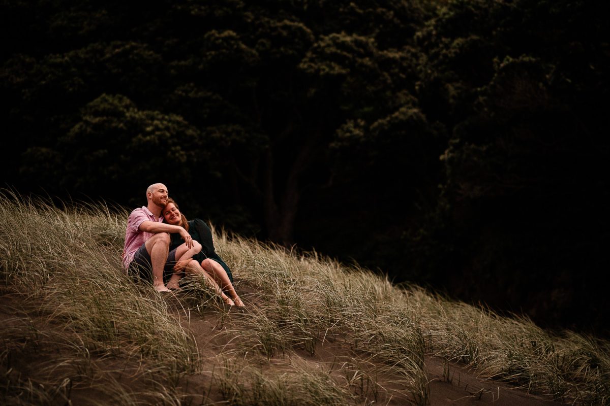 A young couple share an intimate moment on a grassy hill at a picturesque black sand beach near Auckland