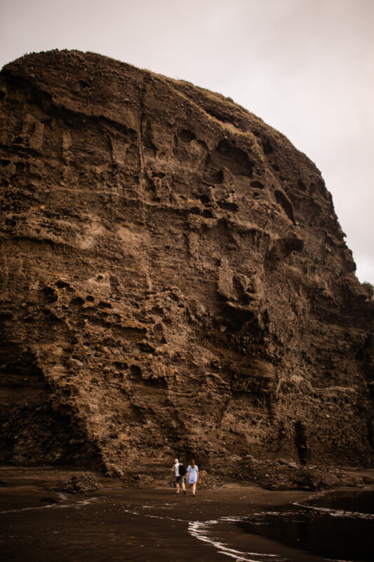 A couple sit under a rock formation at a black sand beach in New Zealand