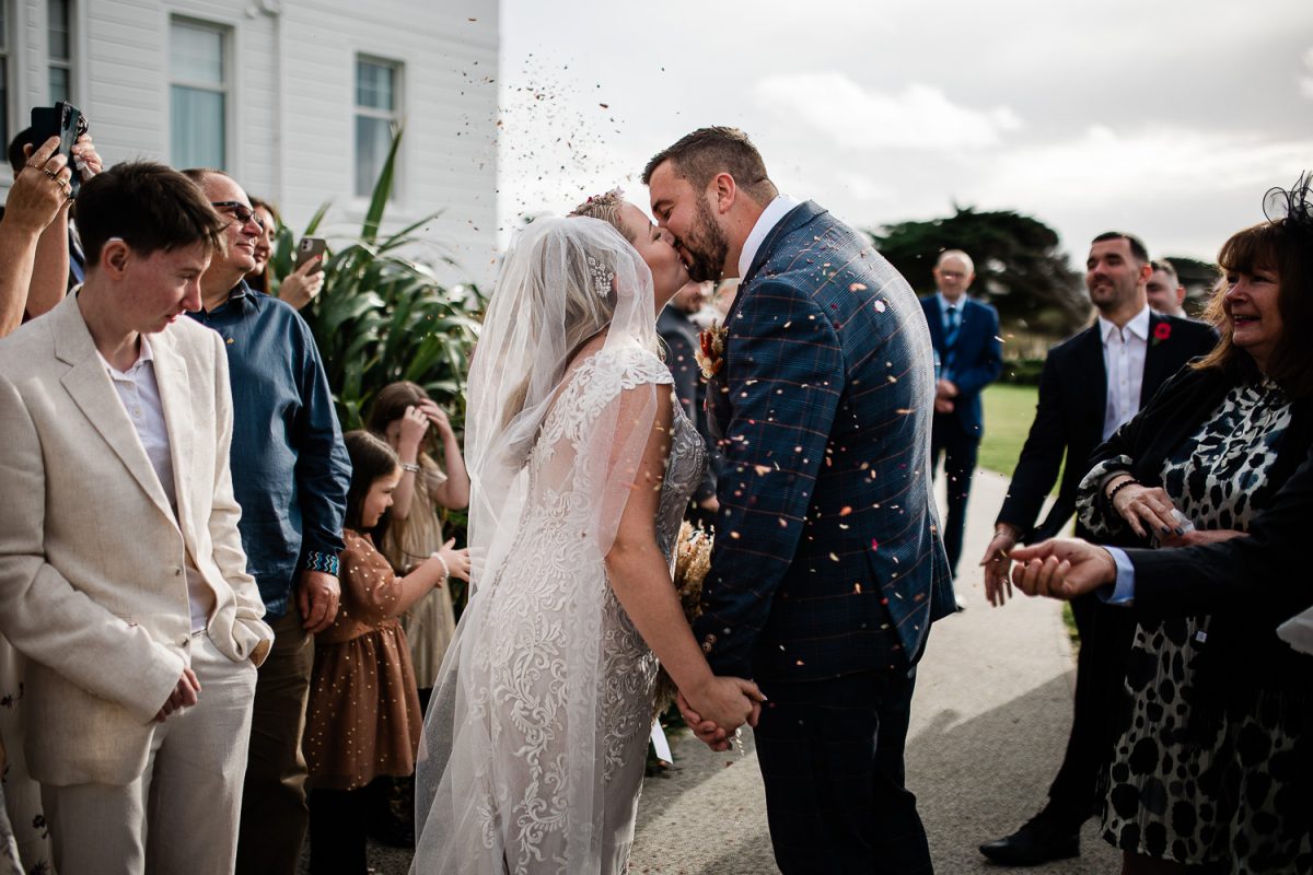 A bride and groom kiss while their friends throw confetti over them for the confetti shot
